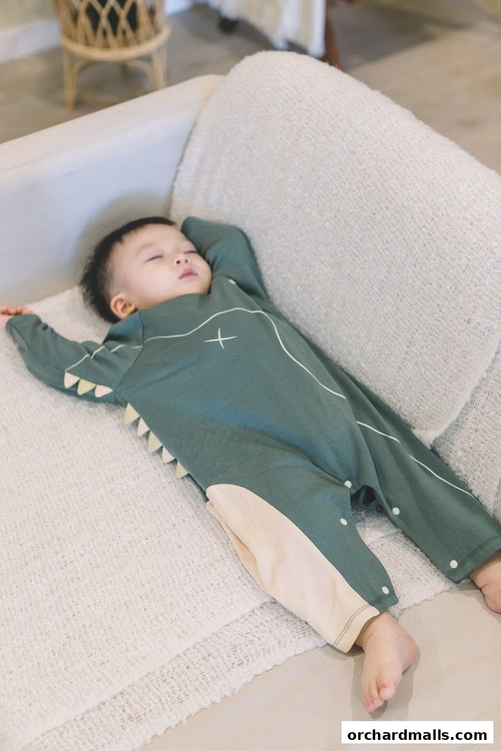A baby sleeps peacefully on a couch wearing a cute dinosaur-themed romper.