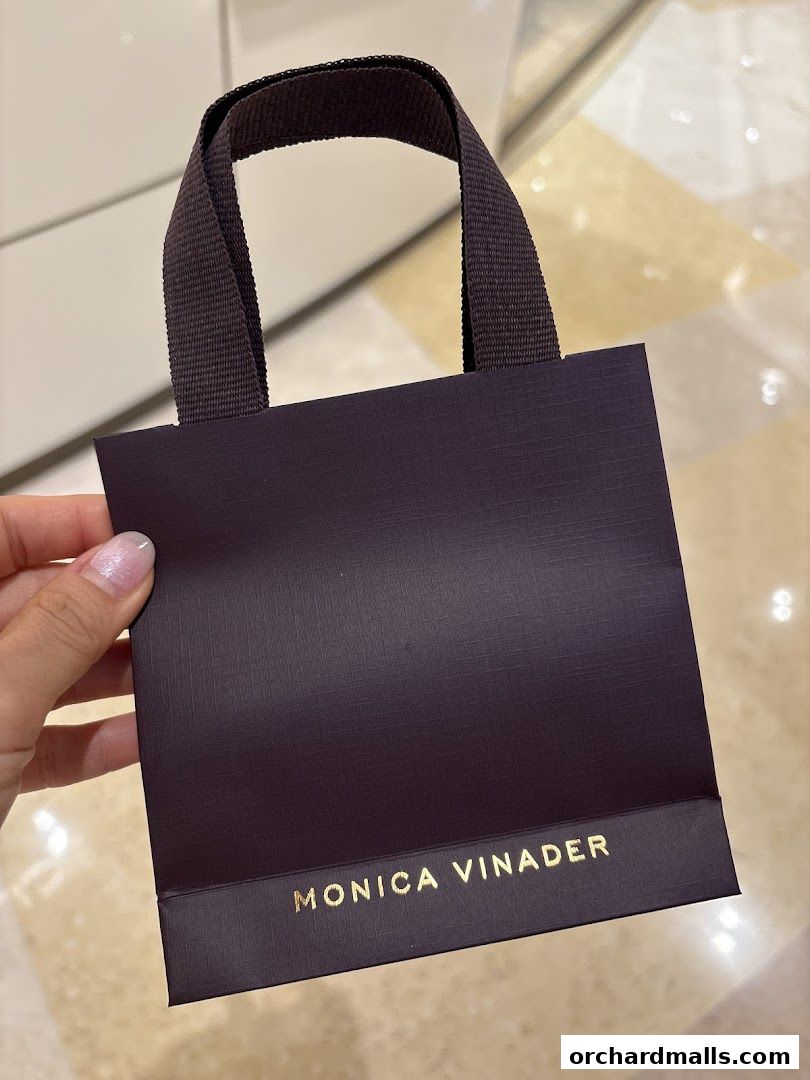 A hand holds a dark purple shopping bag with gold lettering that says Monica Vinader.