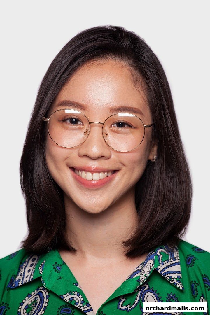 A smiling young woman with dark hair and glasses wears a green patterned shirt.