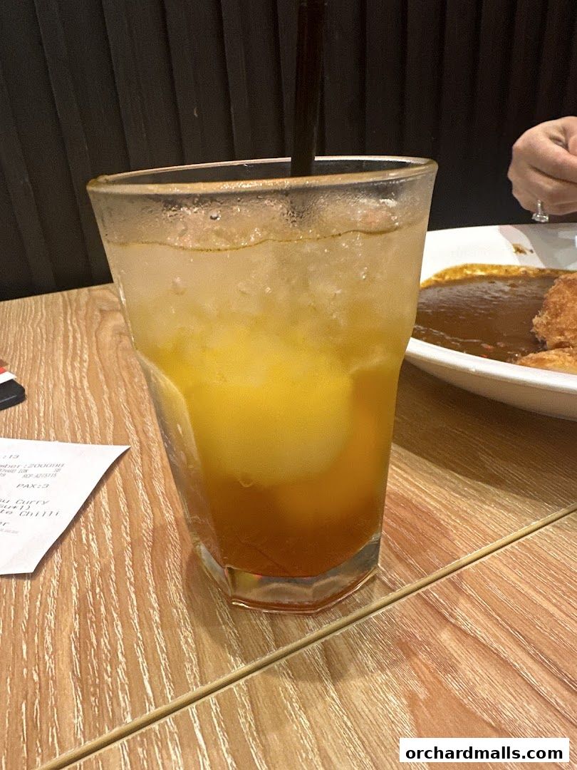 A refreshing beverage with ice and a straw sits on a wooden table next to a plate of curry.
