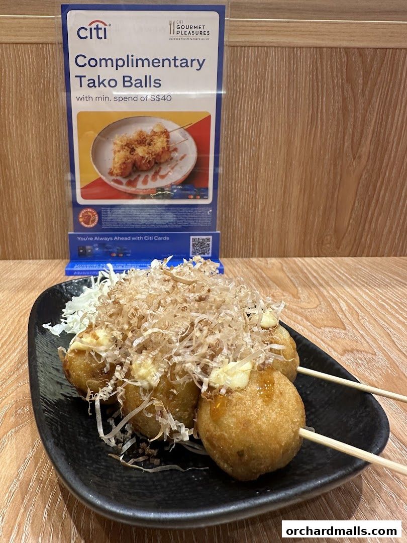 A plate of takoyaki balls topped with bonito flakes and sauce, with a promotional sign in the background.