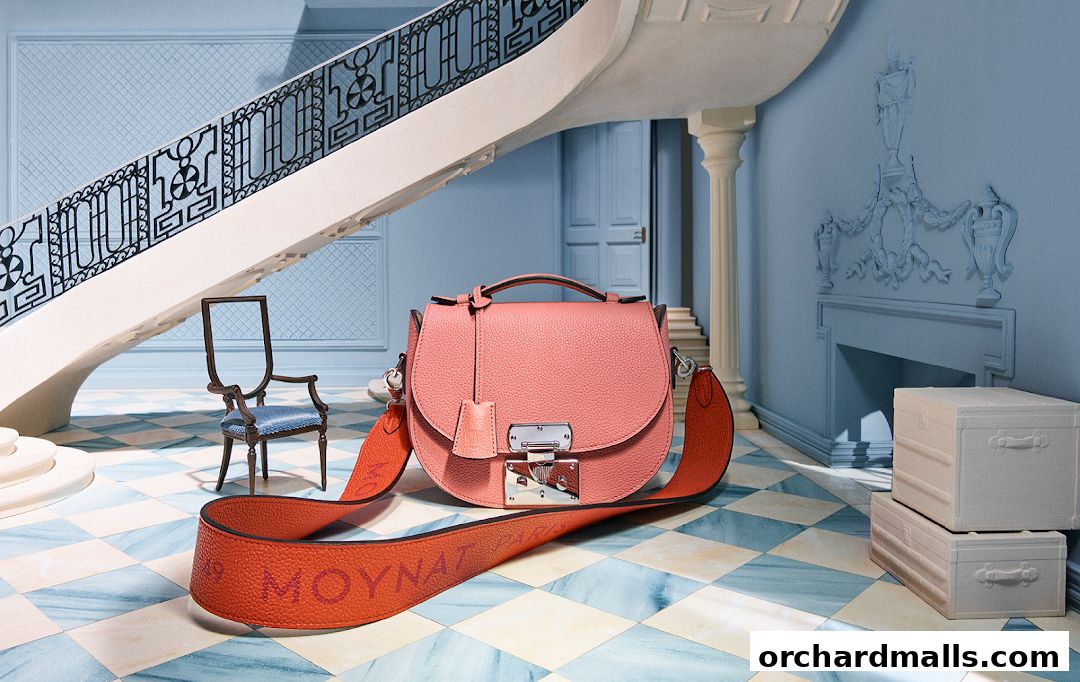 A pink Moynat handbag displayed in an elegant room with a staircase and antique furniture.