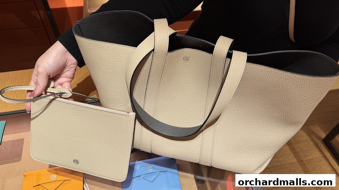 A person holds a beige leather tote bag and matching pouch from Moynat.