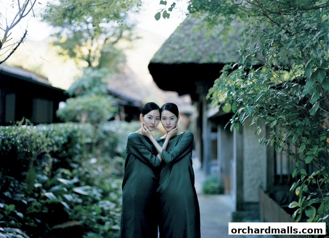 Two women in matching green dresses stand in a serene outdoor setting with traditional architecture.