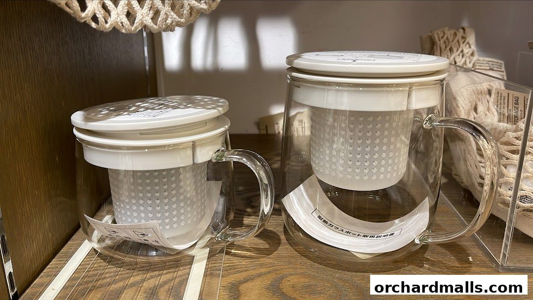 Two clear glass teapots with white infusers are displayed on a wooden shelf.