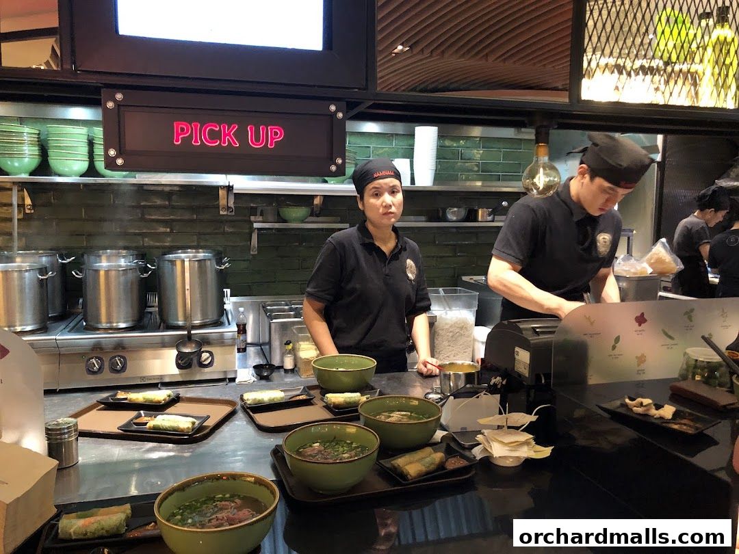 Staff preparing food behind a counter with bowls of soup and spring rolls.