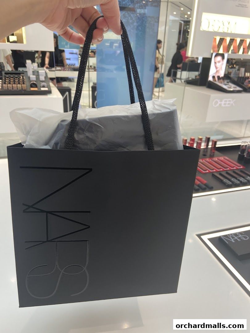 A hand holds a black NARS shopping bag filled with products inside a beauty store.