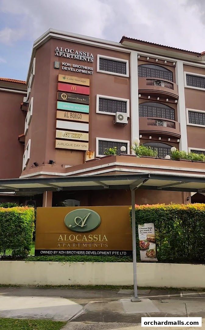 Exterior view of Alocassia Apartments building with various shop signs.