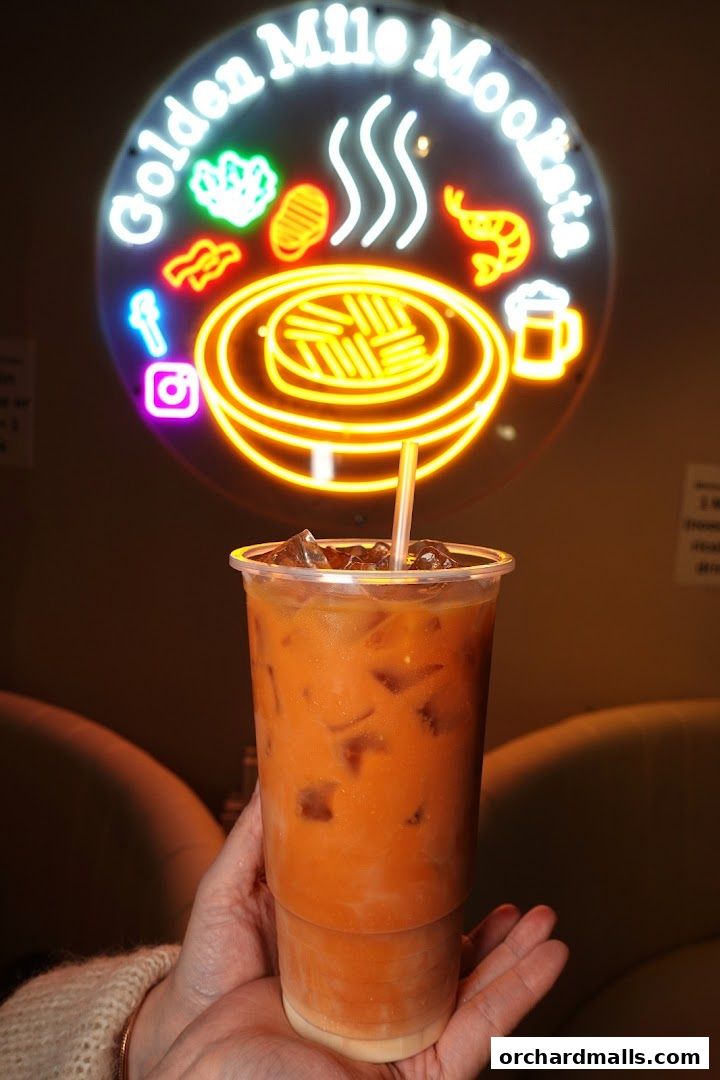 A hand holds a refreshing Thai iced tea in front of a neon sign for Golden Mile Mookata.