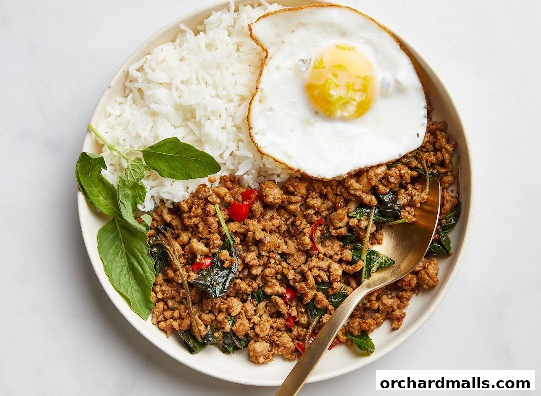 A plate of Thai basil stir-fry with rice and a fried egg, garnished with fresh basil leaves.