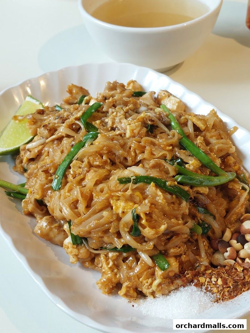 A plate of Pad Thai noodles with chicken, lime, peanuts, and chili flakes, served with a side of broth.