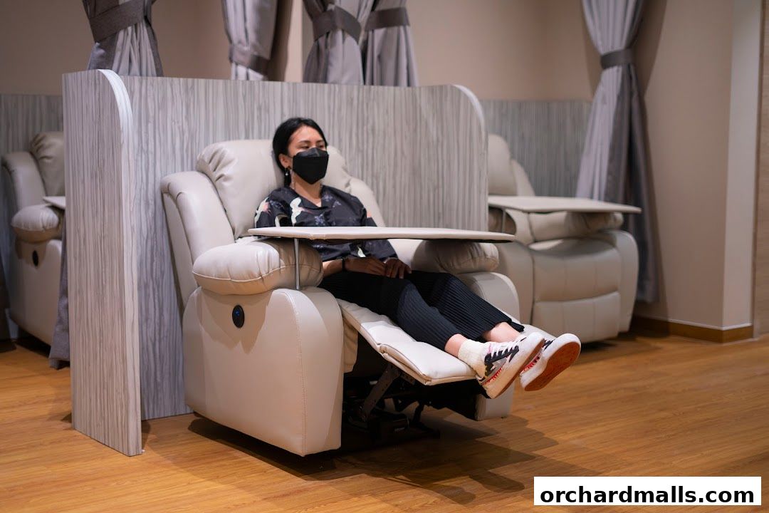 A woman wearing a mask relaxes in a recliner chair with a tray table.