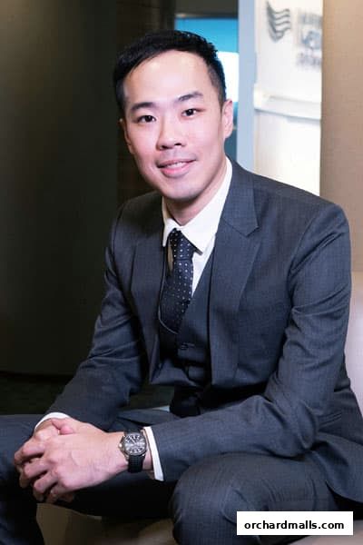 A man in a suit and tie sits with his hands clasped, looking at the camera.