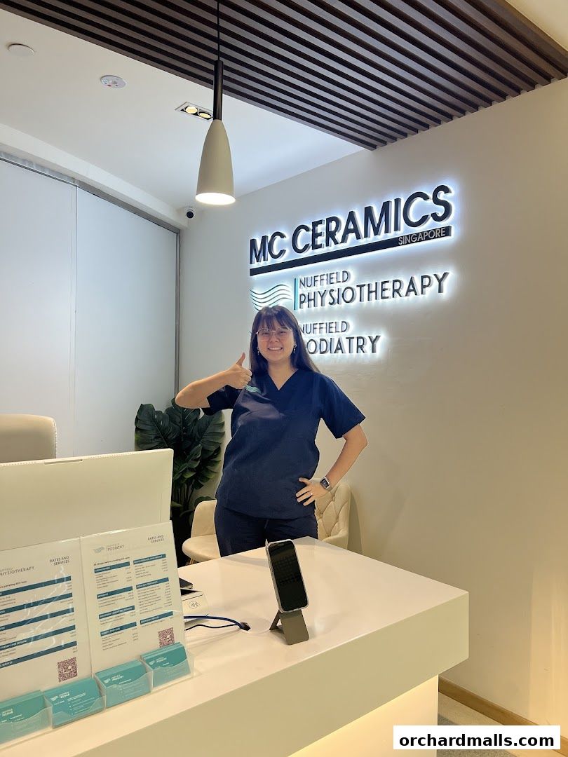 A woman in scrubs gives a thumbs-up at the reception desk of Nuffield Physiotherapy.