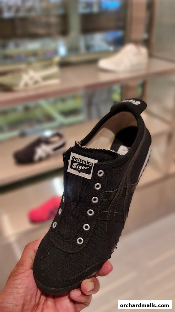 A hand holds a black Onitsuka Tiger shoe with other shoes blurred in the background.