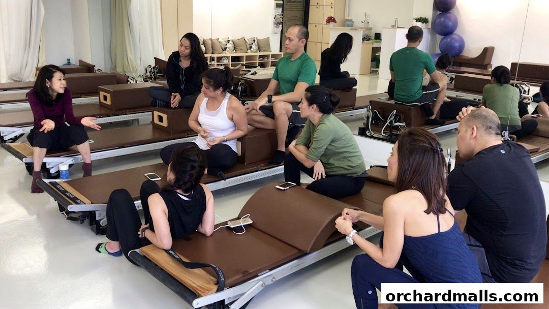 A group of people are sitting on Pilates reformers in a studio, listening to an instructor.