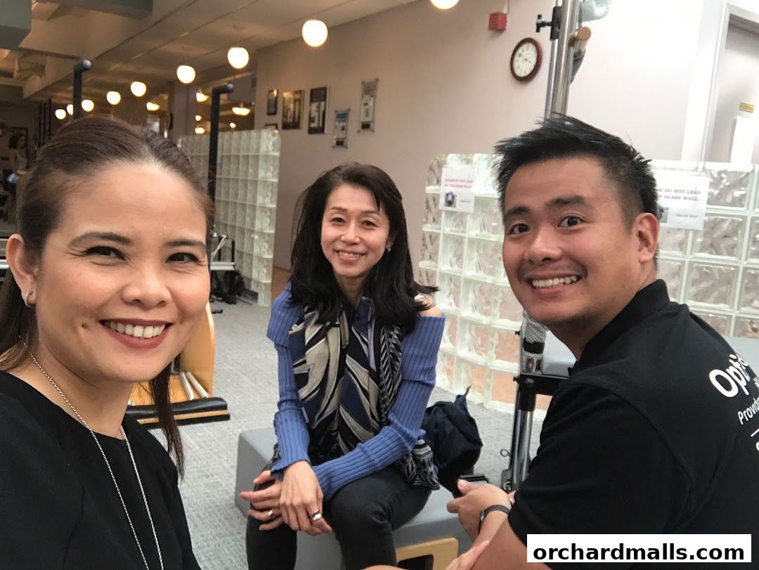 Three smiling people pose for a selfie inside a bright, modern studio with Pilates equipment.