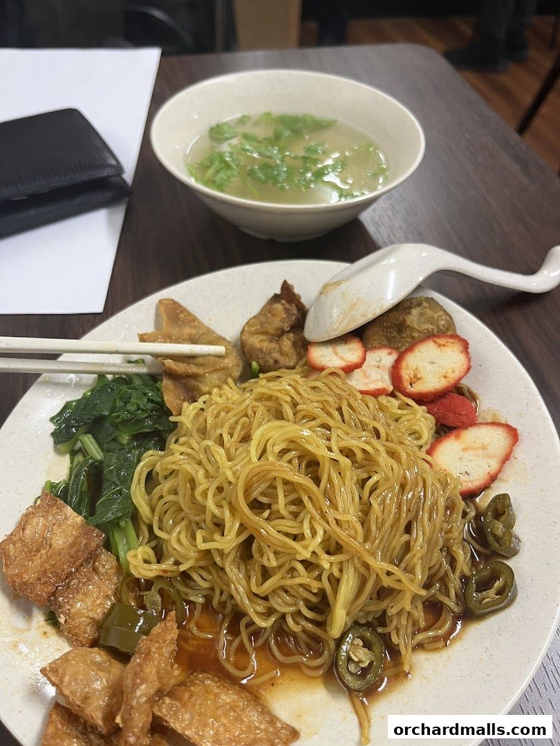 A plate of noodles with soup, vegetables, and fried items, served with chopsticks and a spoon.