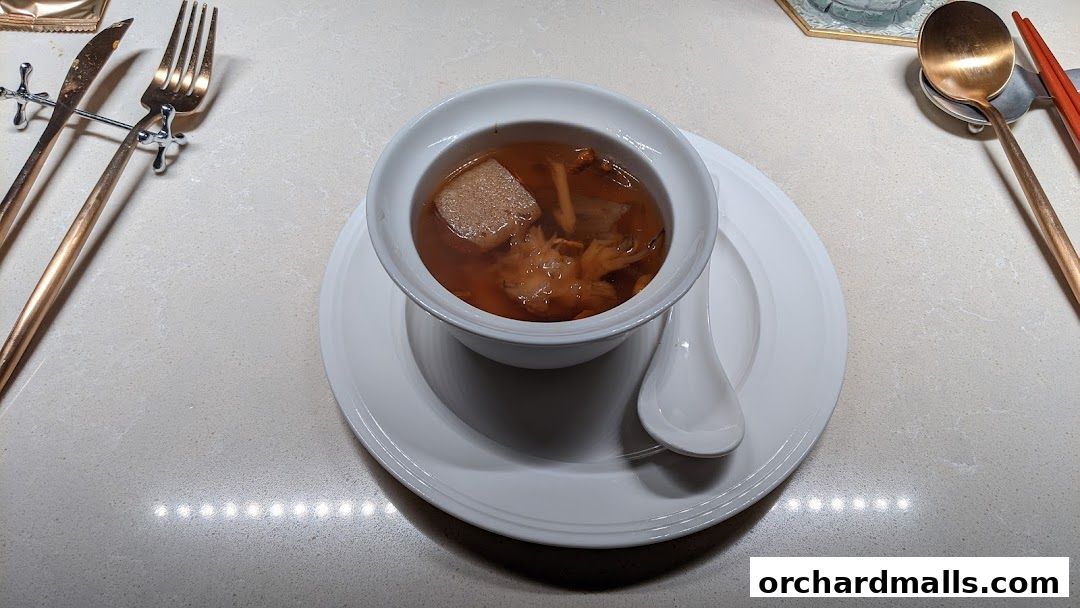 A bowl of rich Chinese soup with mushrooms and a piece of meat, served with cutlery.