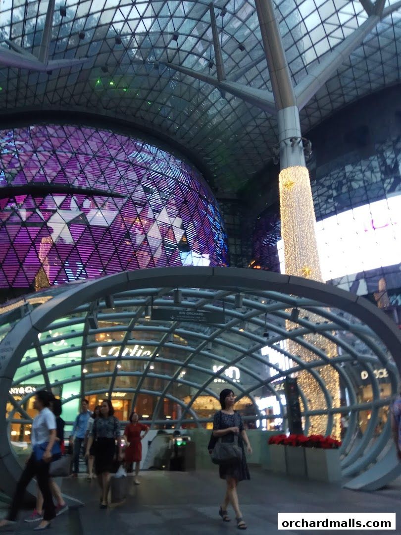 People walking through a modern shopping mall with a distinctive glass dome and illuminated pillar.