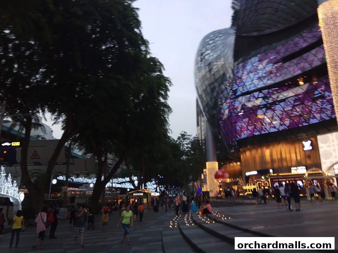 A street scene at dusk with people walking past illuminated shops and trees.