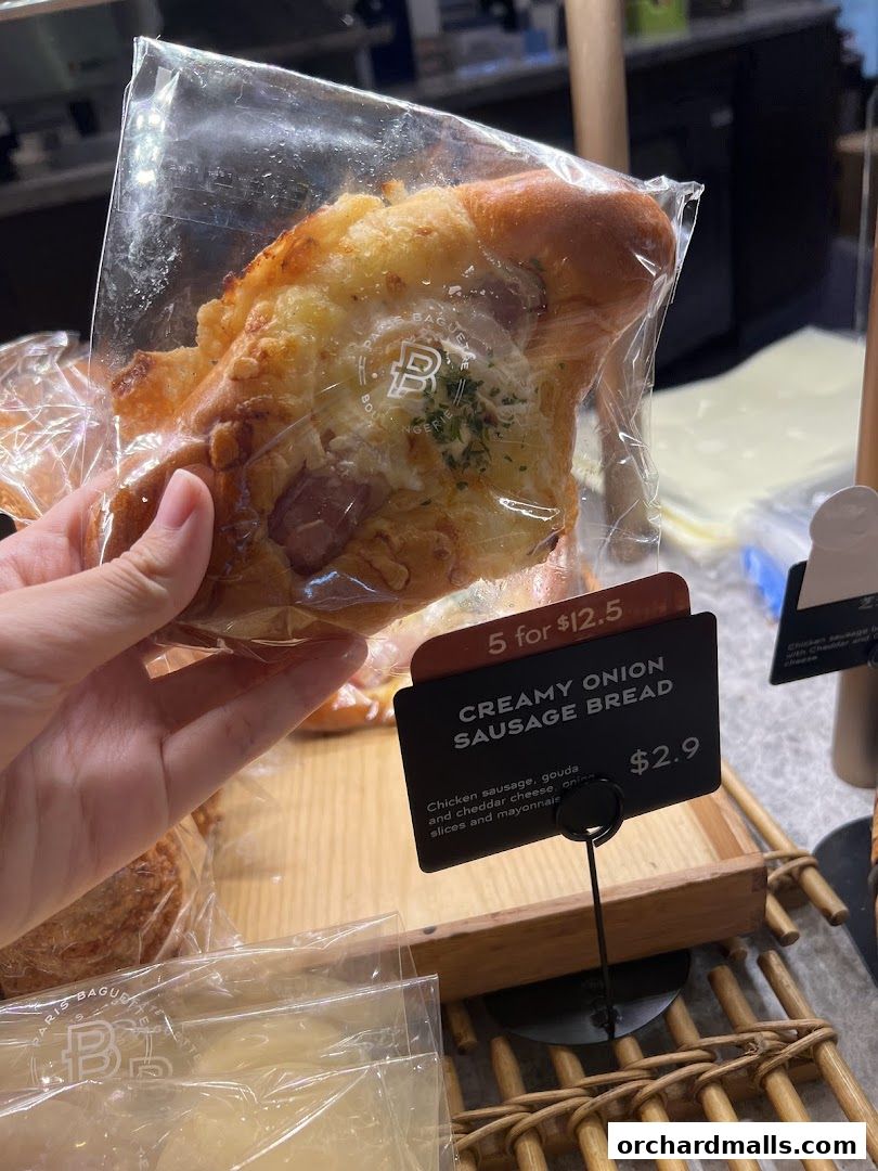A hand holds a packaged creamy onion sausage bread from Paris Baguette.