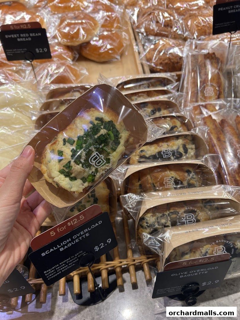 A hand holds a scallion overload baguette in front of a display of various baked goods.