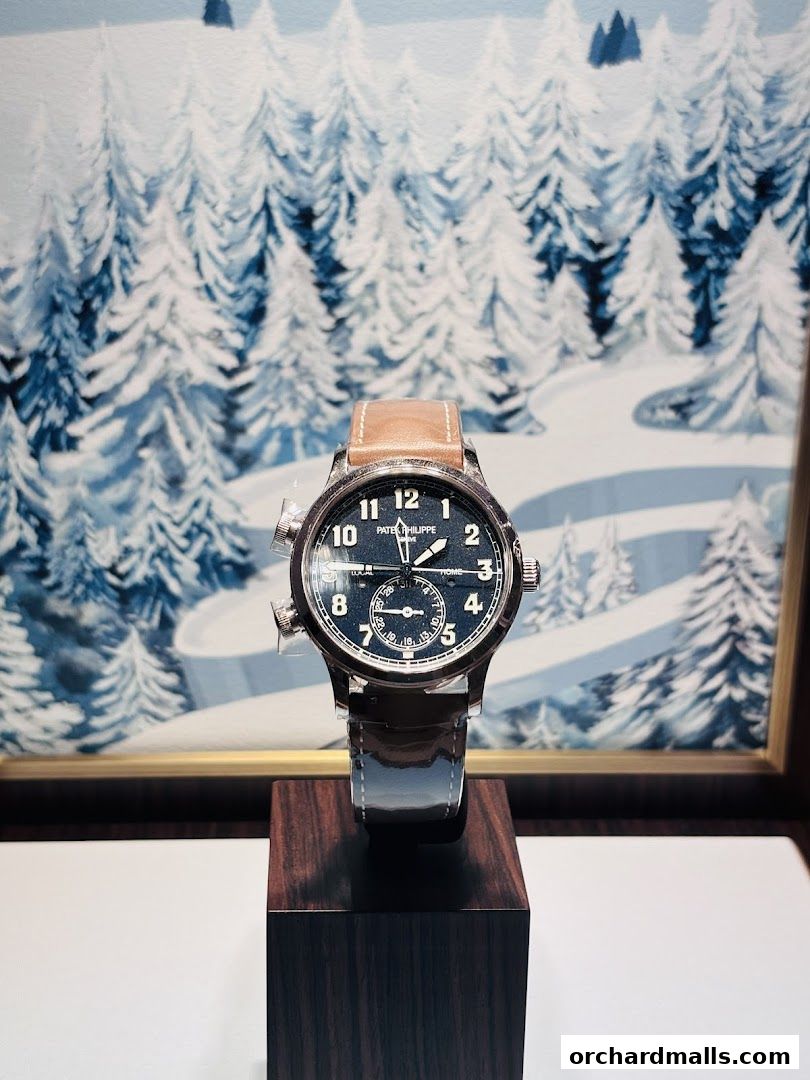 A Patek Philippe watch with a dark dial and leather strap displayed in front of a winter forest painting.