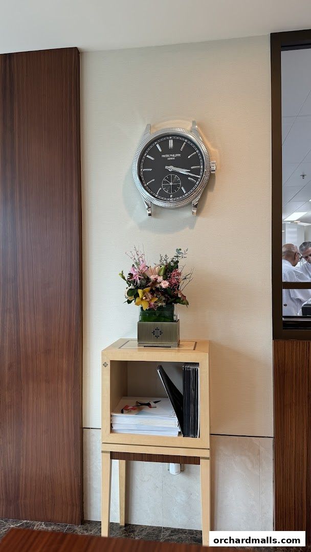 A Patek Philippe branded clock hangs above a display cabinet with flowers and brochures.