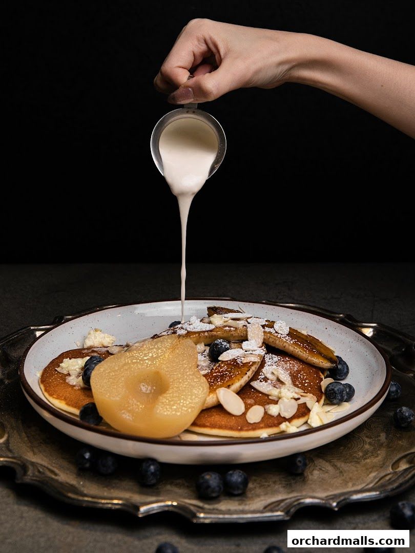 A hand pours cream over a stack of pancakes topped with fruit and nuts.