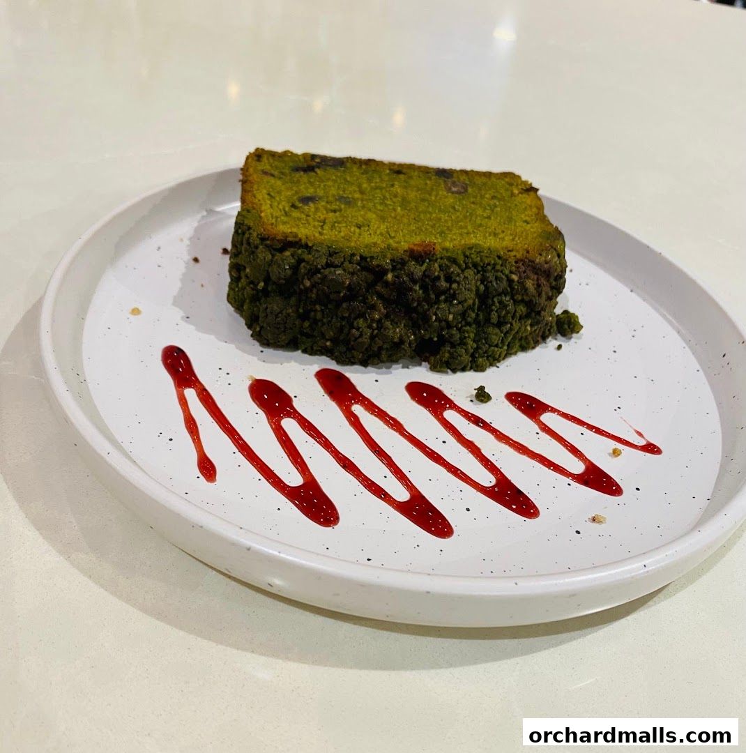 A slice of green tea cake with red berry sauce artfully drizzled on a white plate.