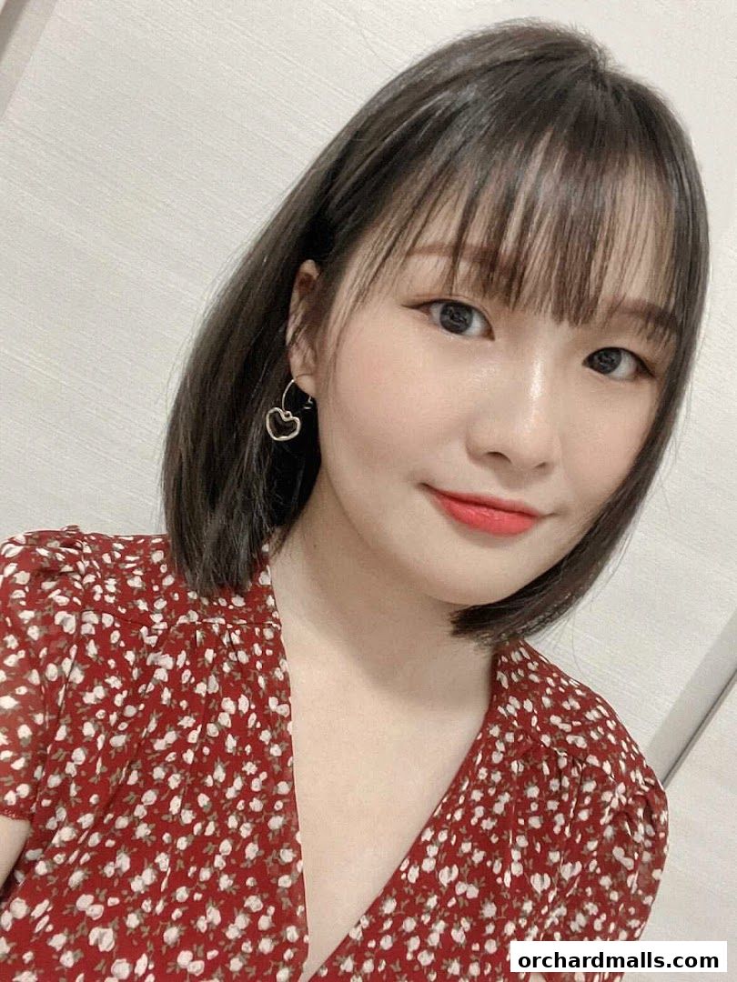 A young woman with short dark hair and bangs wears a red floral dress and cherry earrings.
