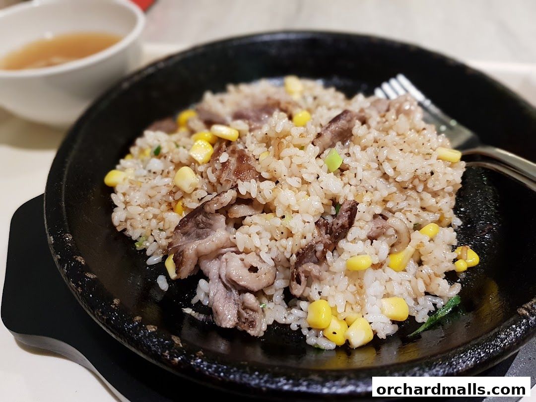 A close-up shot of a sizzling plate of fried rice with beef and corn, served with a side of broth.