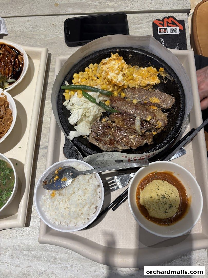 A sizzling steak platter with corn, scrambled eggs, and vegetables, served with rice and mashed potatoes.