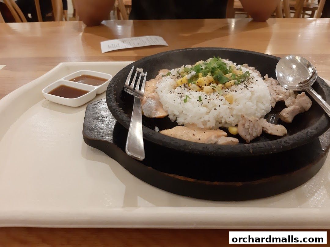 A sizzling hot plate meal with rice, meat, corn, and sauce, served with a fork and spoon.