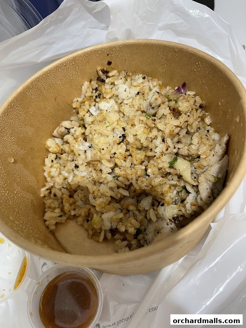 A close-up of a takeaway bowl filled with fried rice and chicken, with a side of sauce.