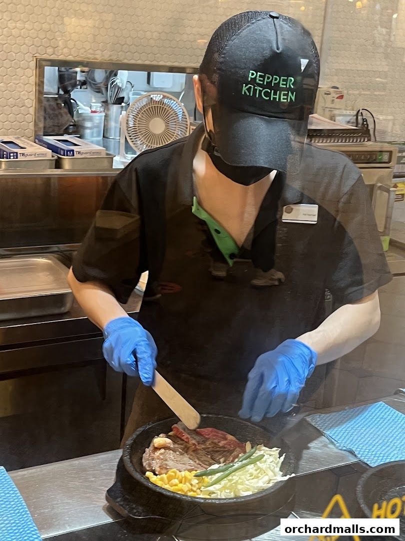 A chef in a black cap and apron prepares a sizzling stone bowl dish with steak, corn, and vegetables.