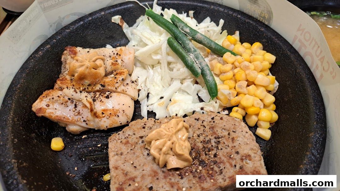 A sizzling hot plate with grilled chicken, steak, corn, green beans, and shredded cabbage.