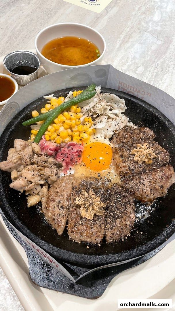 A sizzling hot plate meal with steak, chicken, egg, corn, and beans, served with soup.