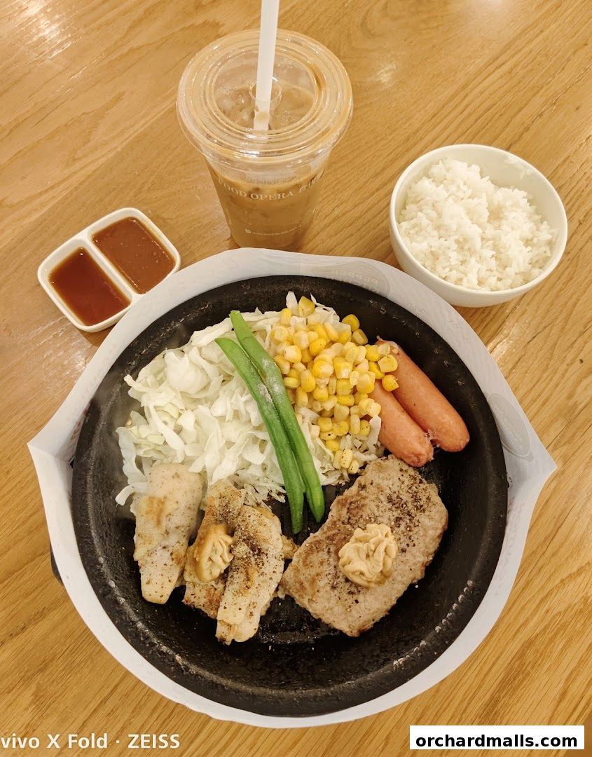 A sizzling hot plate meal with steak, chicken, sausages, corn, beans, cabbage, rice, and a drink.