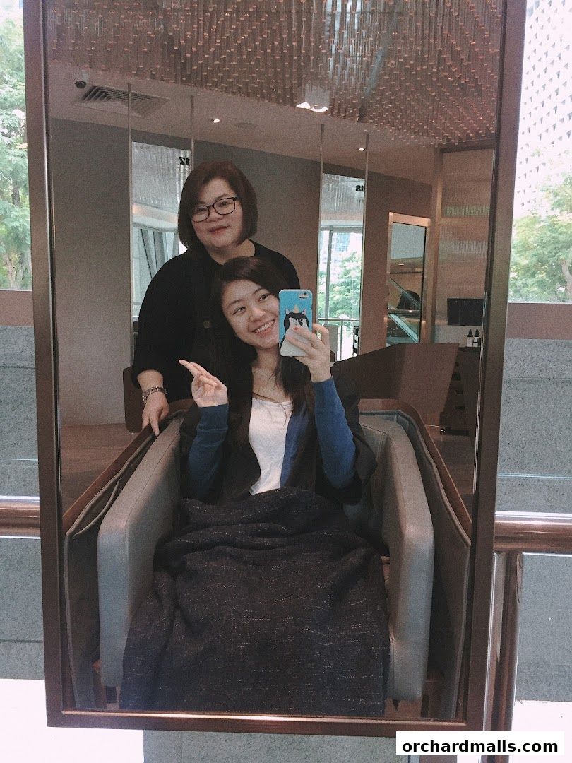 A woman takes a selfie with a stylist in a salon chair, reflecting in a mirror.
