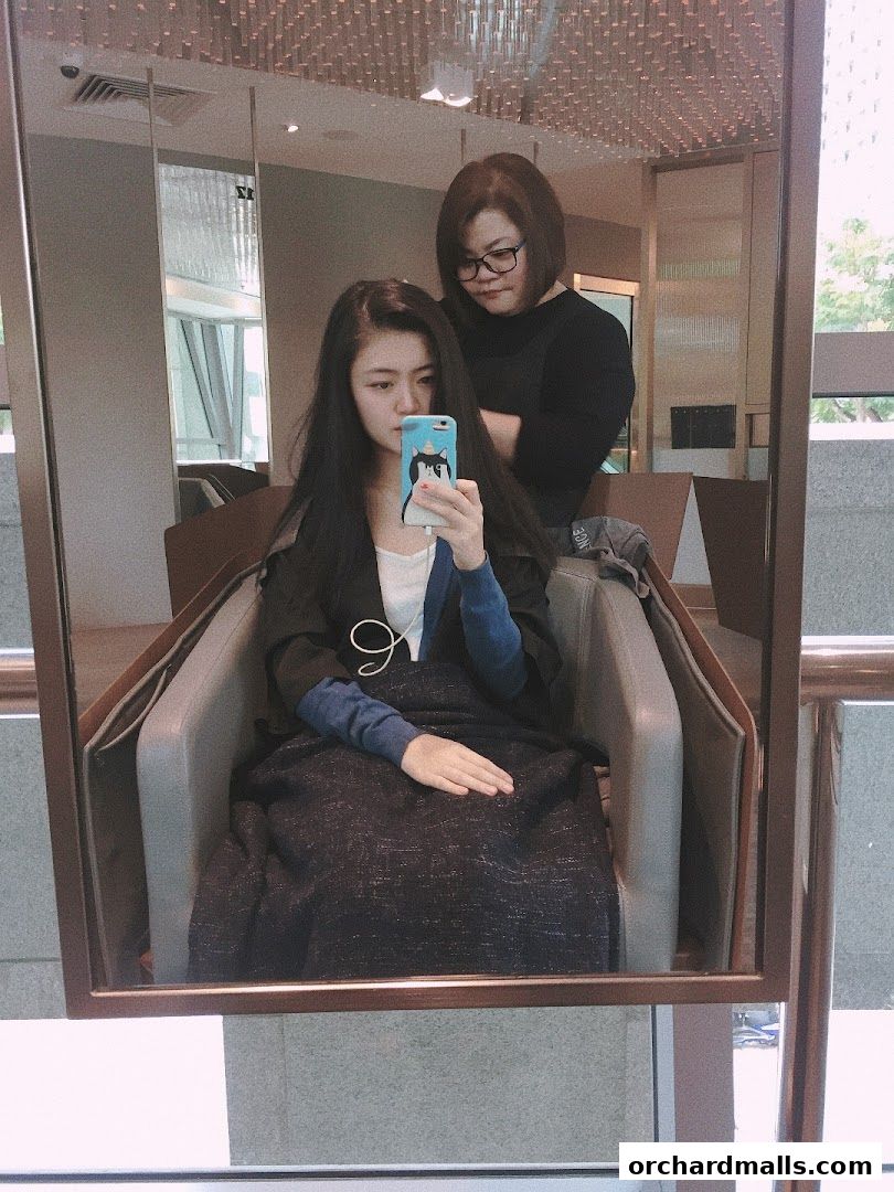 A woman takes a selfie in a salon chair while a stylist works on her hair.