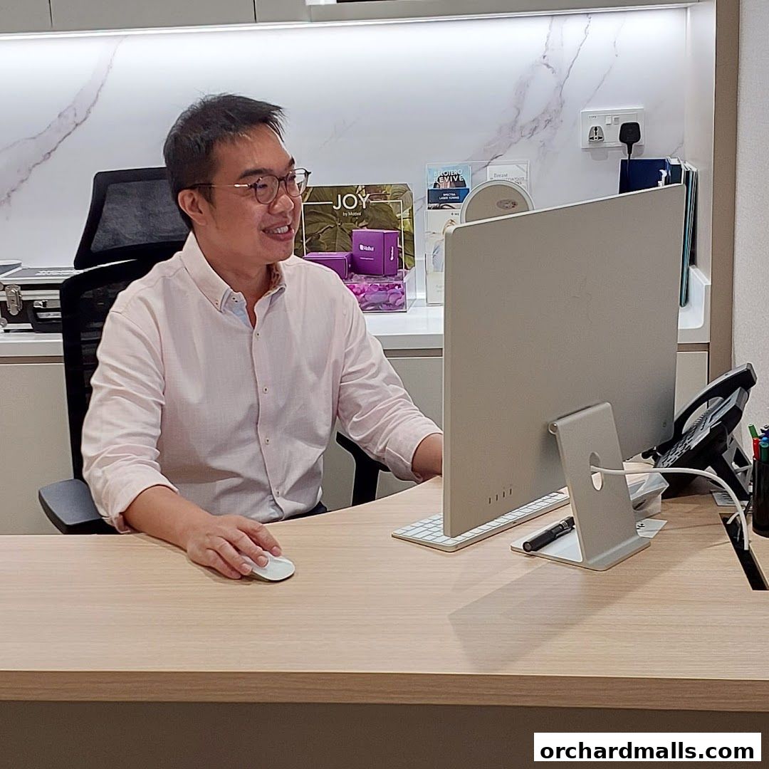 A man in a pink shirt sits at a desk in front of a computer at Picasso Plastic Surgery.