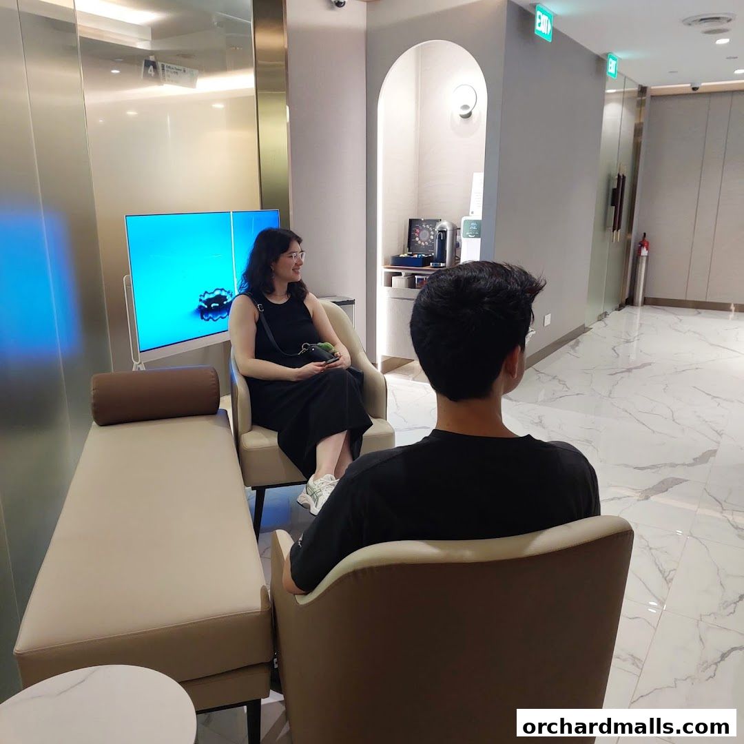 People waiting in a modern, clean reception area with a TV screen.