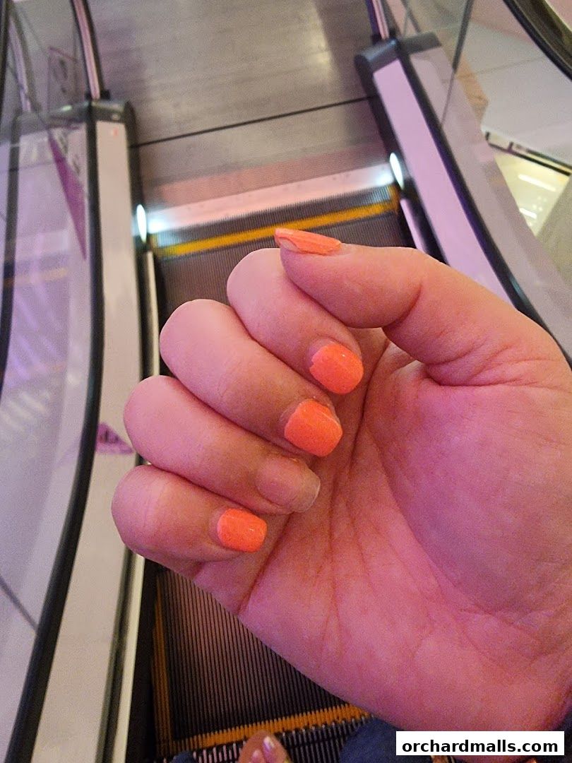 A close-up of a hand with bright orange nails, with escalators in the background.