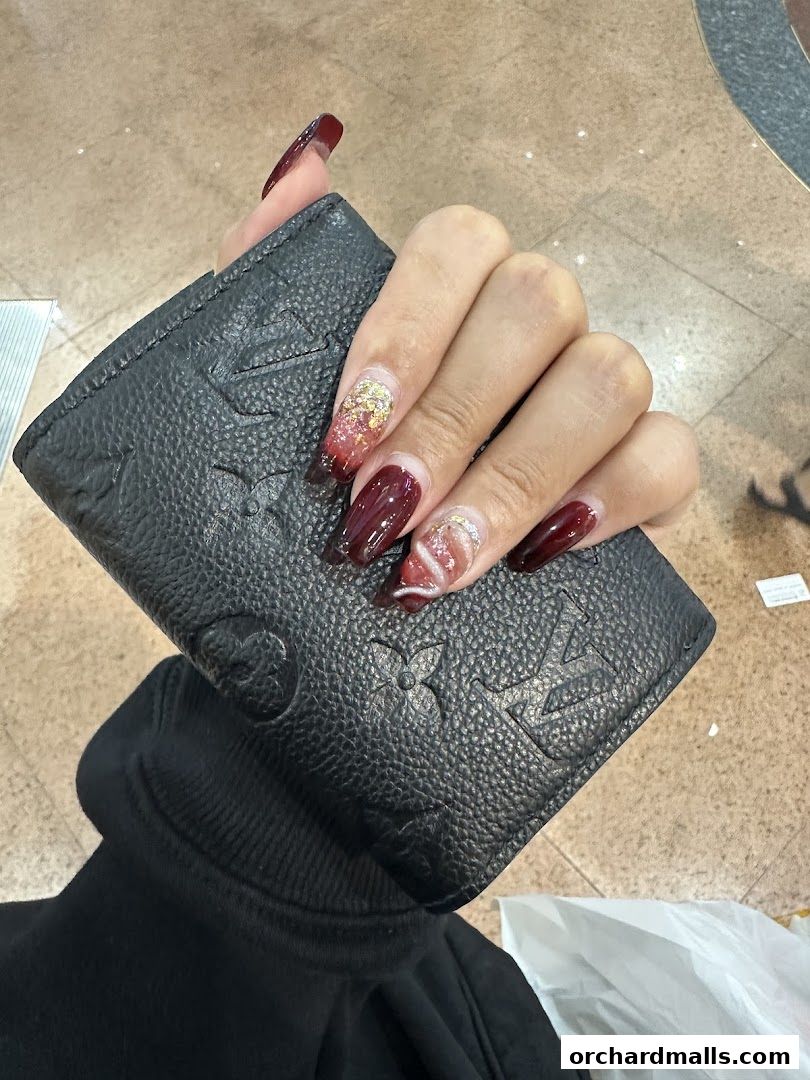 A hand with stylish burgundy and gold glitter nails holds a black textured wallet.