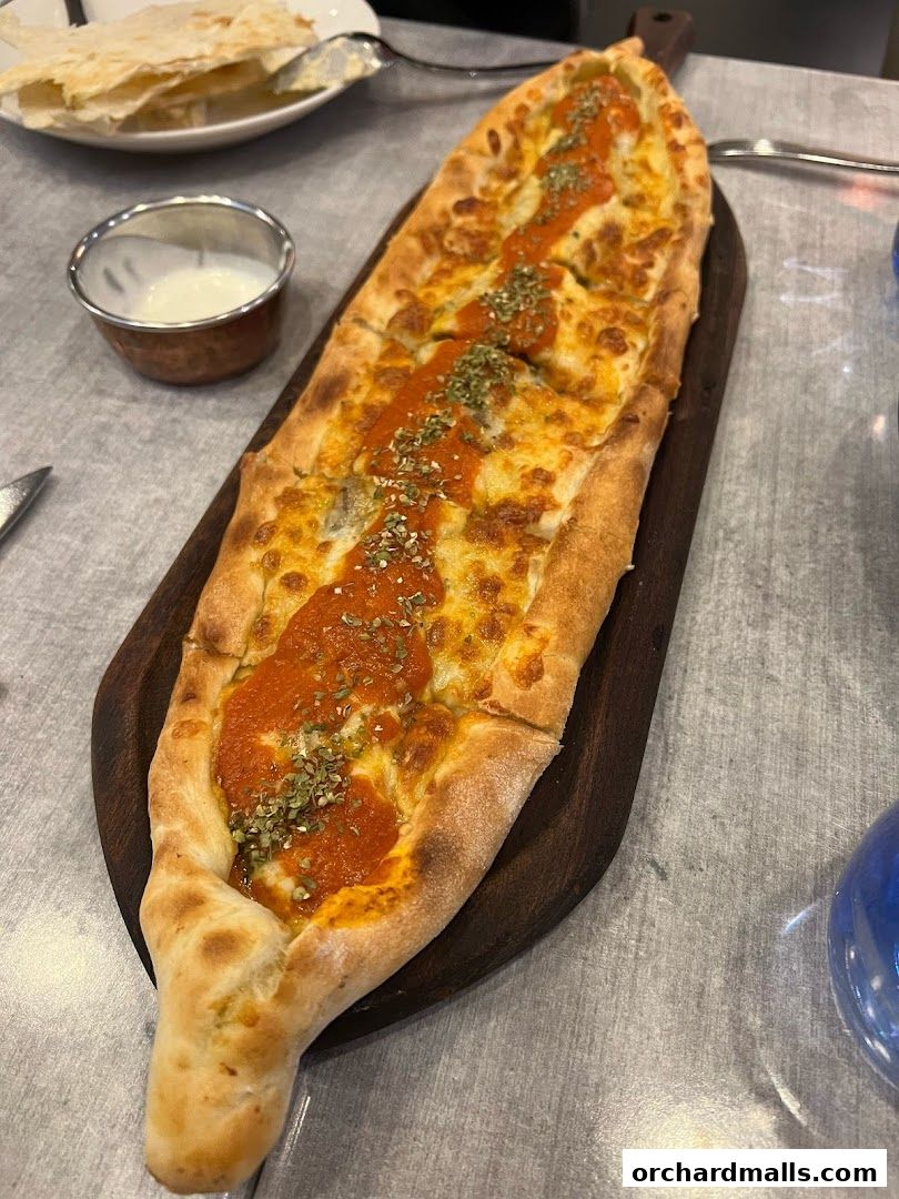 A long, boat-shaped flatbread topped with cheese and tomato sauce, served with a side of dip.