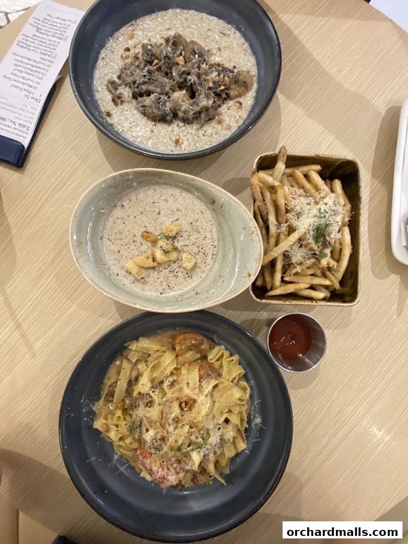 A top-down view of a table with three dishes: pasta, mushroom risotto, and fries, along with a small cup of ketchup.