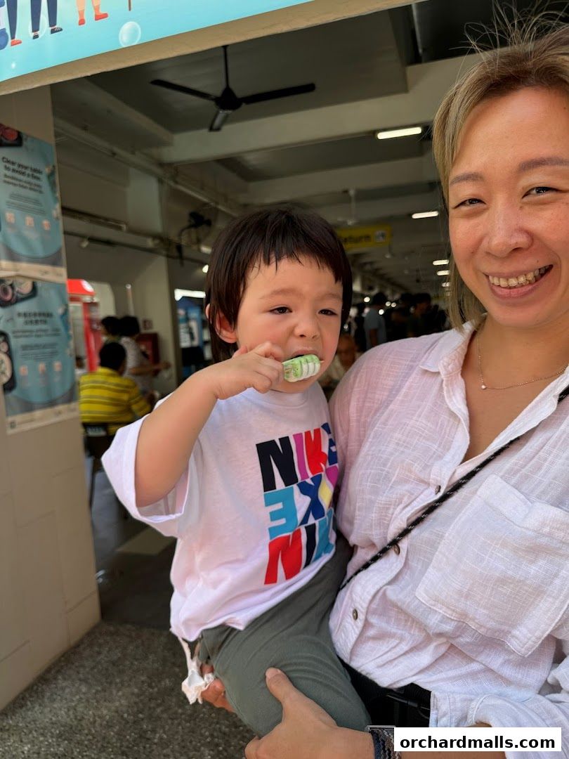 A woman holds a child eating a popsicle in front of a shop.