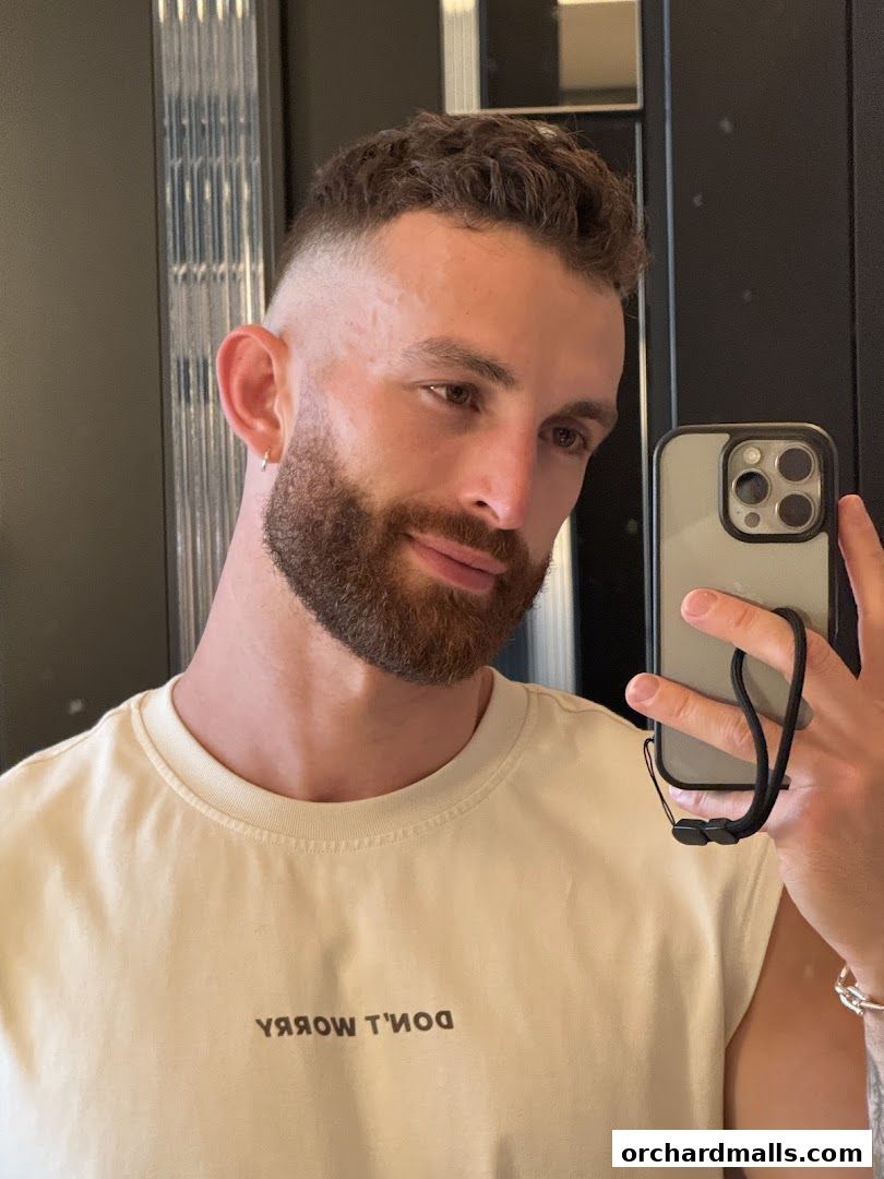 A man with a beard and fade haircut takes a selfie in a mirror.